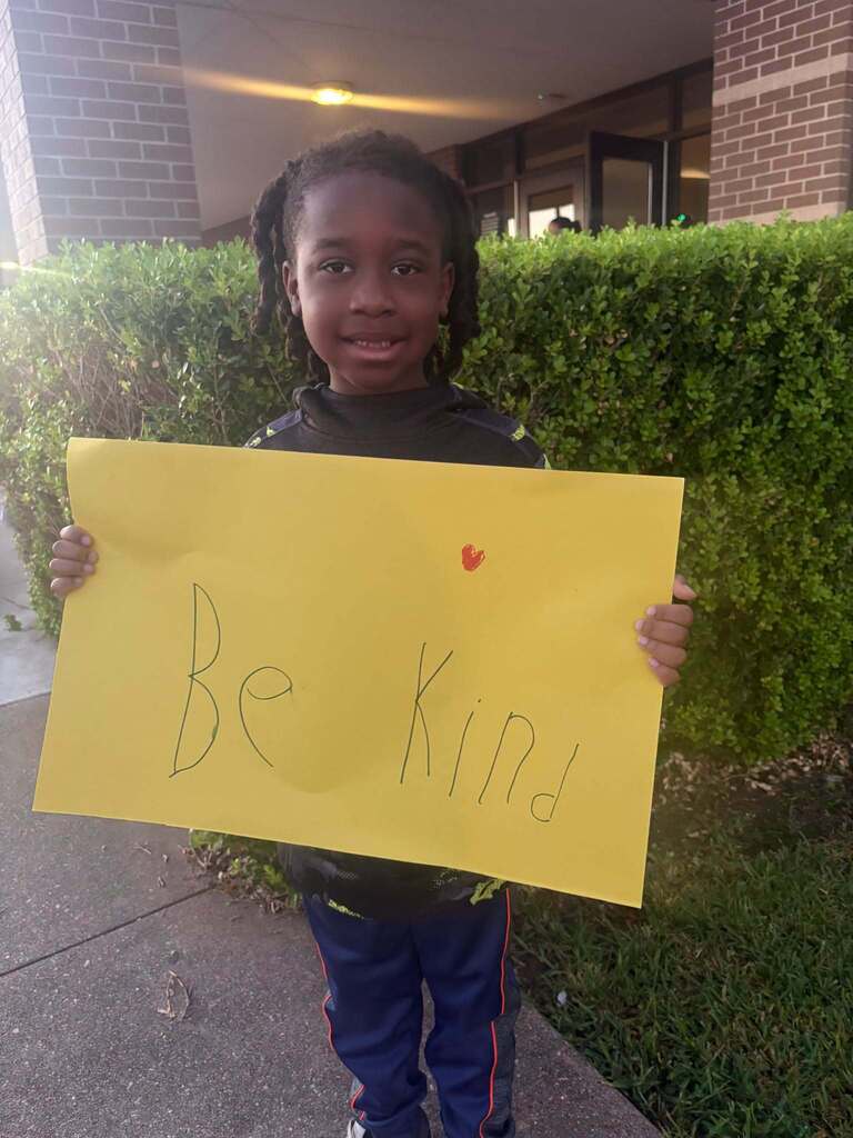 A child holds a yellow sign with the message "Be Kind." Green hedges and a building are in the background.
