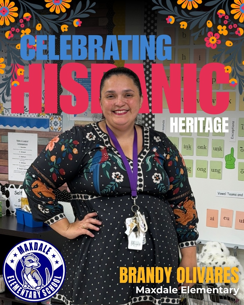 A smiling woman stands in a classroom with vibrant decorations, wearing a colorful floral dress and a purple lanyard.