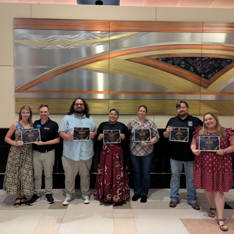 Group of people in formal and casual clothing holding awards in front of a large wall.