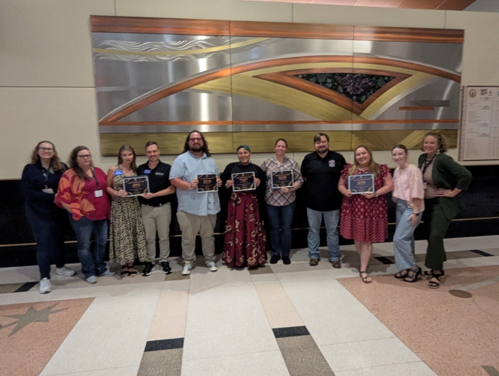 A group of people stands in a hallway, holding awards, with a large metallic art piece on the wall.