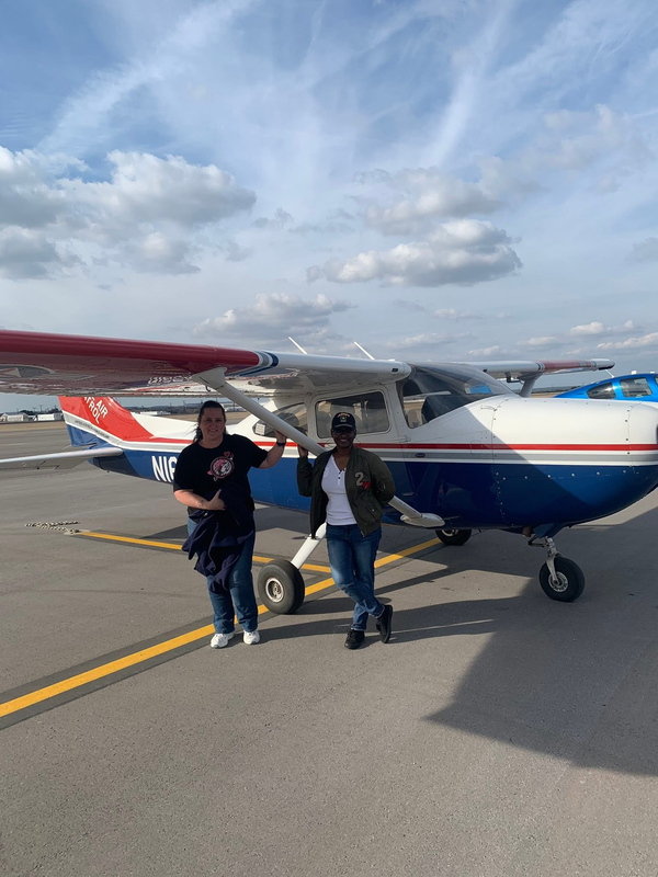 two women standing next to a plane smiling outside
