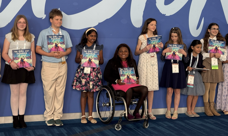 group of students smiling with a book in their hand around a woman in a wheel char