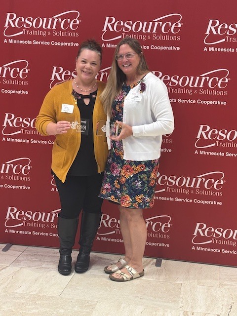 Two women stand side-by-side smiling and holding awards in front of a red backdrop printed with the logo "Resource Training & Solutions: A Minnesota Service Cooperative." The woman on the left wears a mustard yellow cardigan over a black top, black pants, and black boots. The woman on the right wears a floral print dress under a white cardigan with metallic sandals. Both are holding clear glass or acrylic recognition plaques.