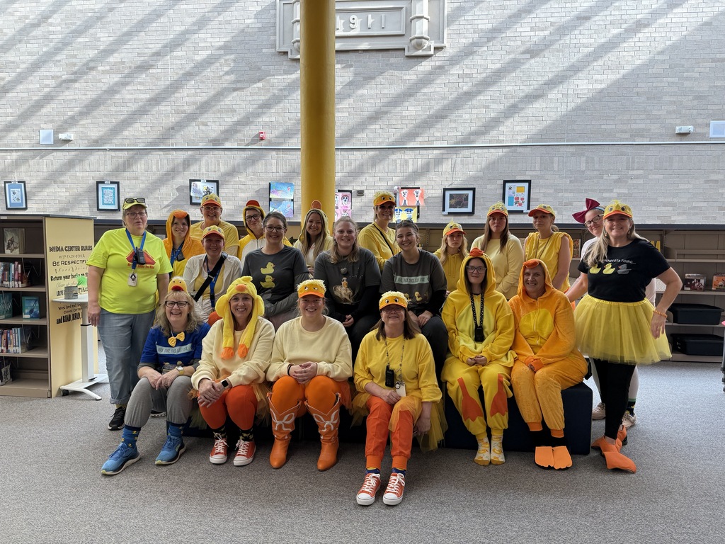 A group of elementary school staff members dressed up in yellow and orange duck costumes and themed t-shirts for Halloween, smiling together while sitting and standing in a row in front of a school library display.