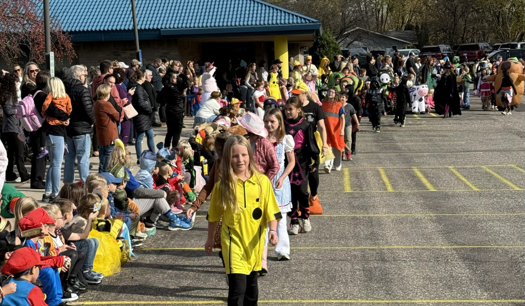 Children in colorful costumes participate in a Halloween parade outdoors, walking in a line past a crowd of families and spectators seated and standing along the sidelines. A young girl in a bright yellow costume leads the procession, with a school building and parked vehicles in the background on a sunny autumn day.