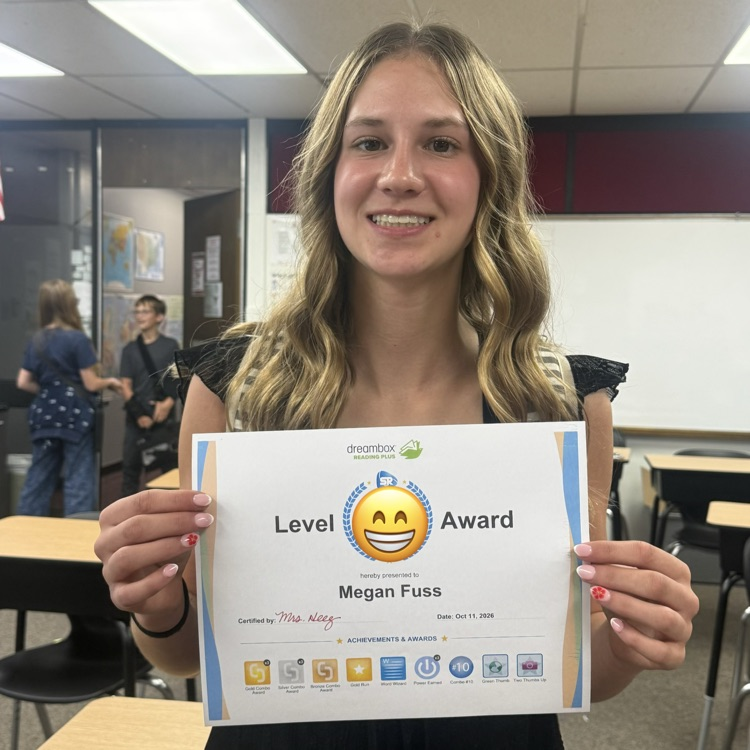 Woman holds up certificate with smiley face award in a classroom with tables and chairs.