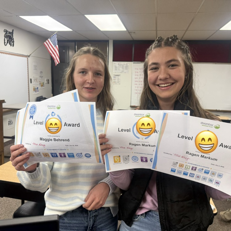 Two young girls with smiley face awards, standing in a classroom, smiling and holding up certificates.