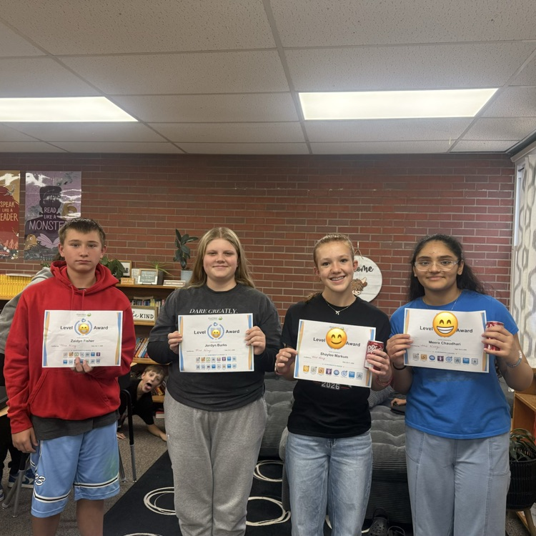 Four students stand in a room with brick walls. Each holds a certificate. A bookshelf and plant are in background.