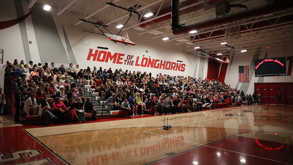 An indoor basketball court with many people sitting on the bleachers, red and white walls, and a large sign.