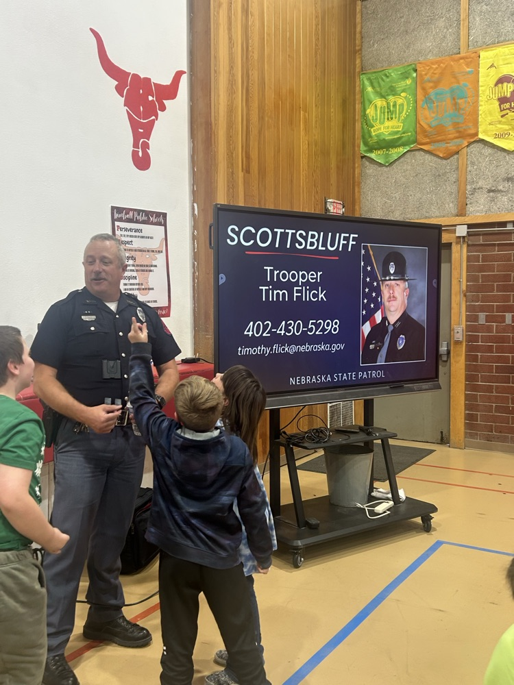 A police officer speaks into a microphone while a boy raises his hand. A screen shows a police officer's photo, name, and phone number.