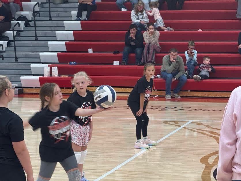 Three young girls in black T-shirts and white shoes play volleyball in a gym with red bleachers.