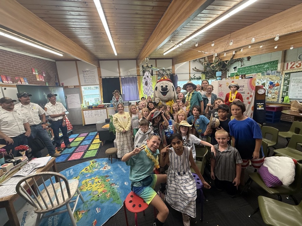 Group of children standing and sitting in a room with a mascot, carpet, and tables.