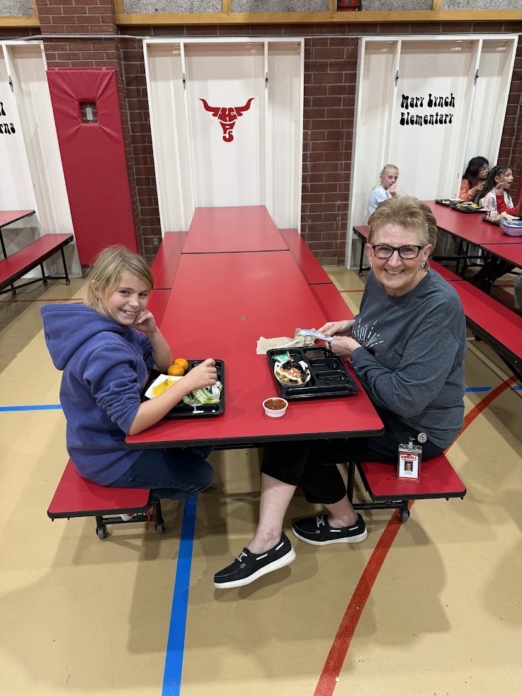 Two people sit at a long red table, each with a plate of food