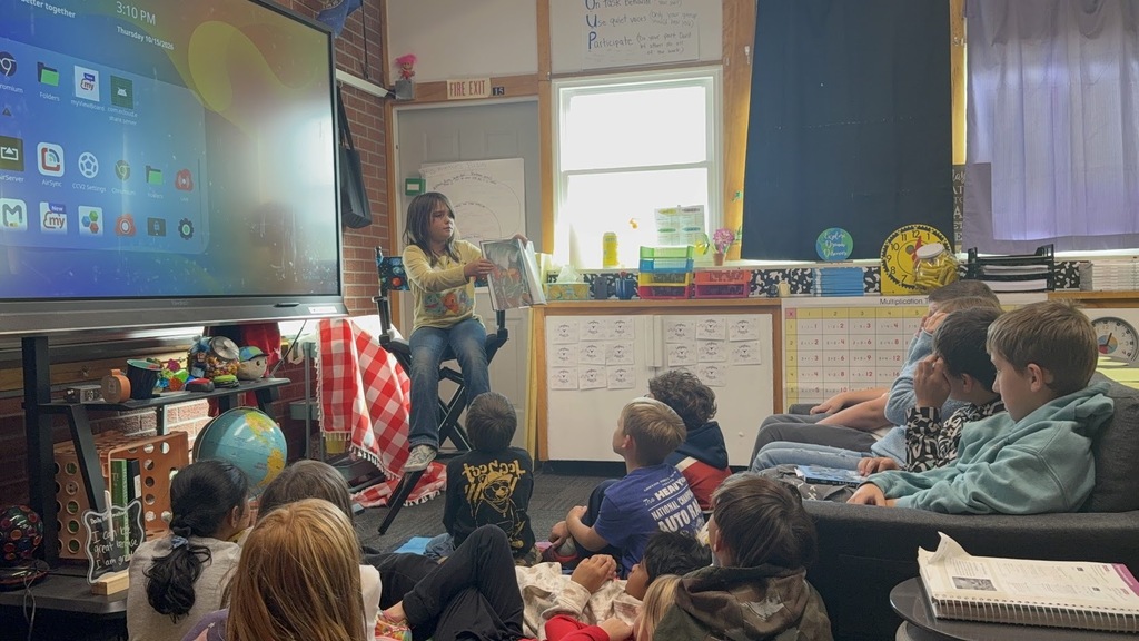 Several children sit on the floor watching a student read a book