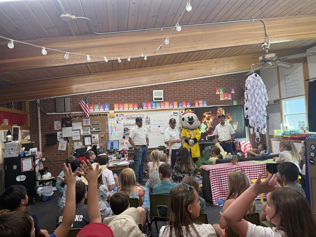 People gathered in a classroom; some seated, some standing; a mascot with yellow and white costume stands with adults.