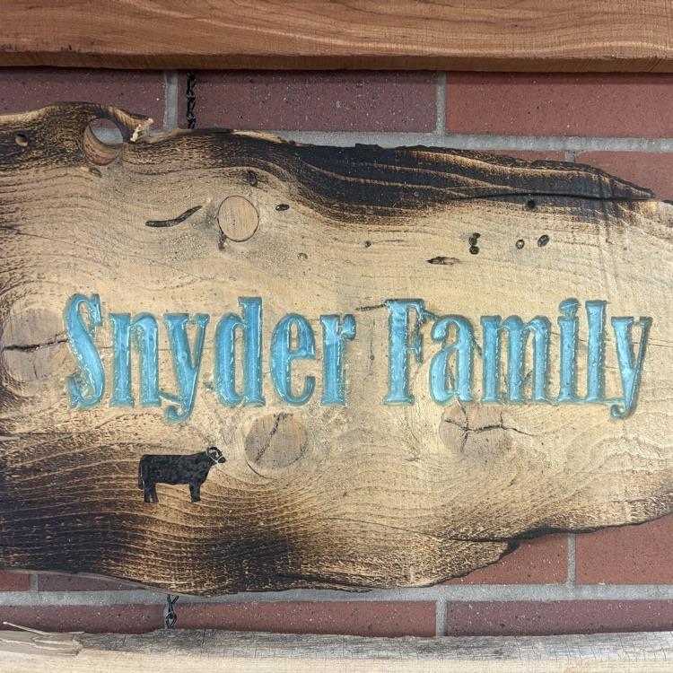 Wooden sign with blue text "Snyder Family" hangs on brick wall with image of a cow.
