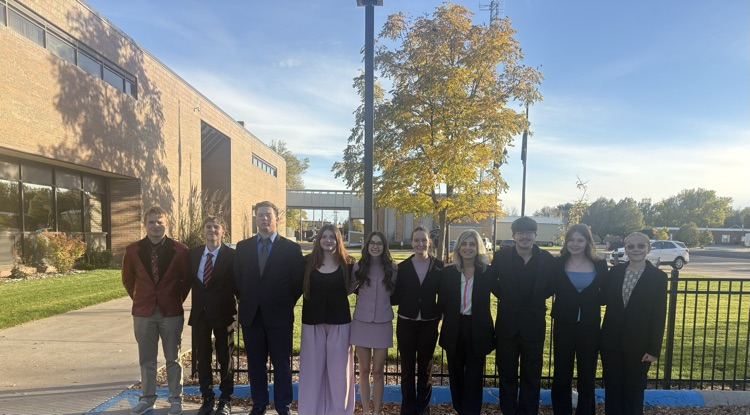 Nine students in suits and dresses standing outside a building, with a fence and tree behind them.