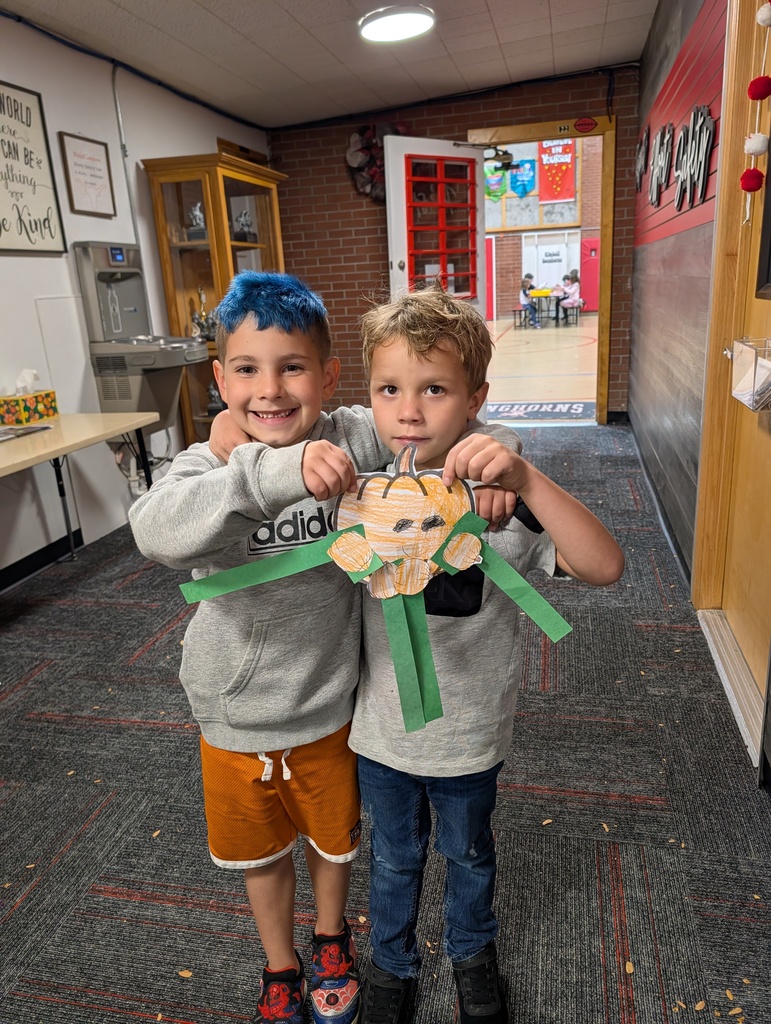 Two young boys stand holding a paper pumpkin with green ribbon, posing for a photo.