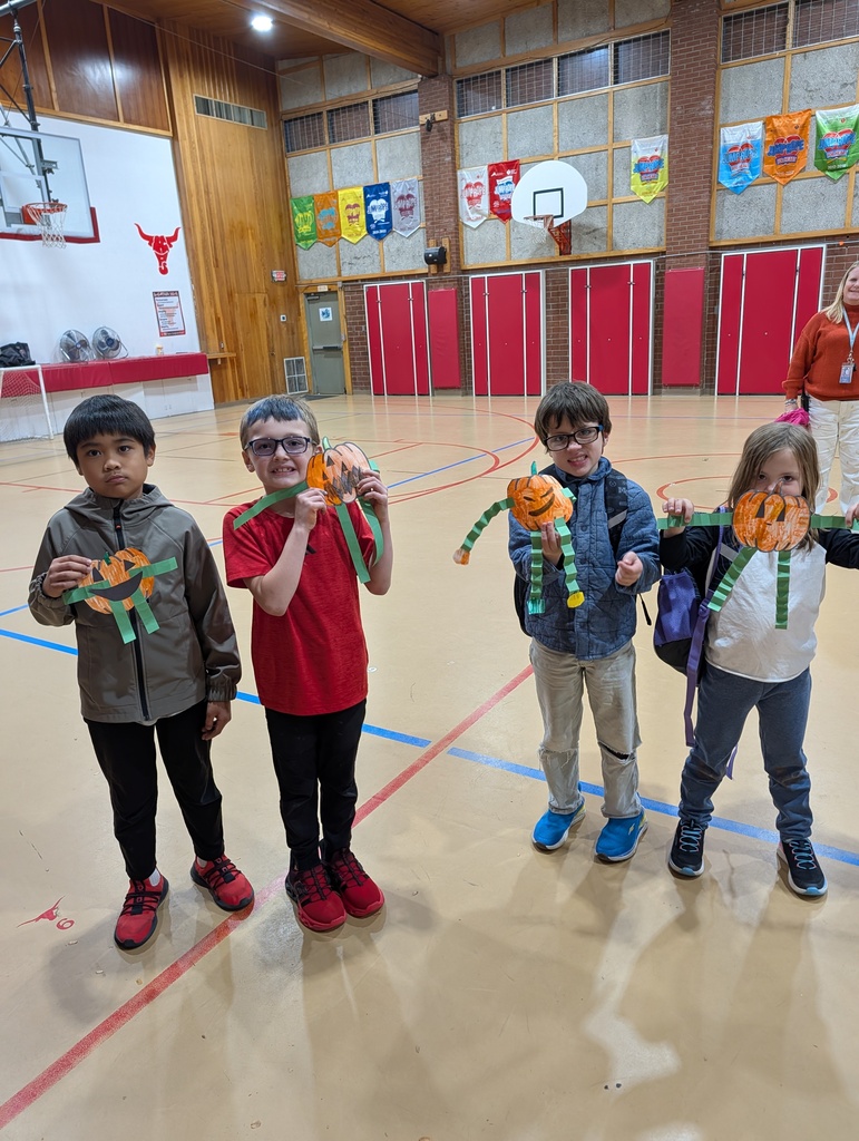 Four students pose in a school gym with paper crafts in their hands. Behind them, a basketball court is visible.