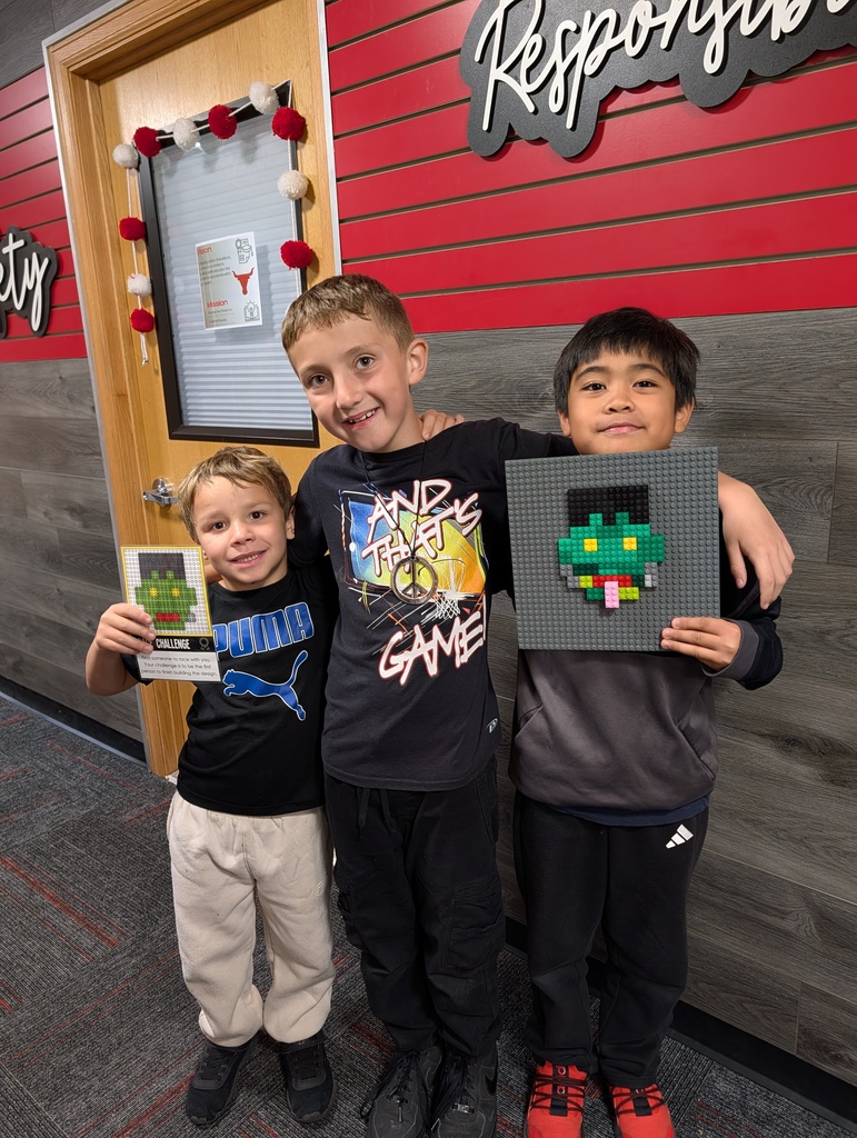 Three boys stand, two holding pixel art, one holding a card, in front of a red wall.