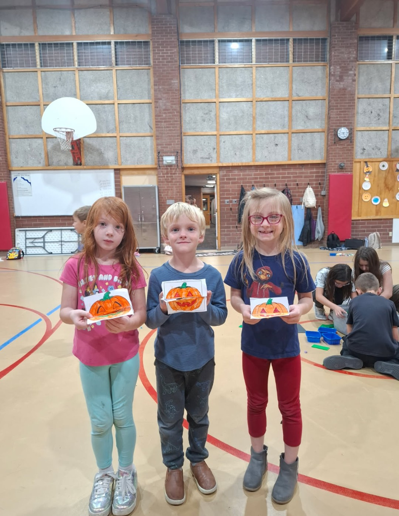 Three children stand in a gym with pumpkin crafts. Two girls and one boy, holding orange pumpkin crafts.