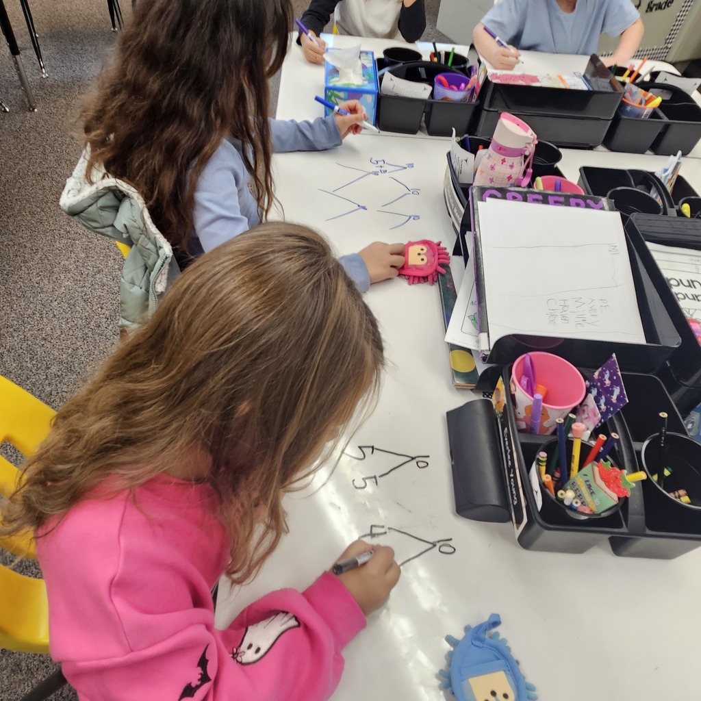 Two young girls draw on a large sheet of paper with markers, one drawing lines and the other drawing shapes.