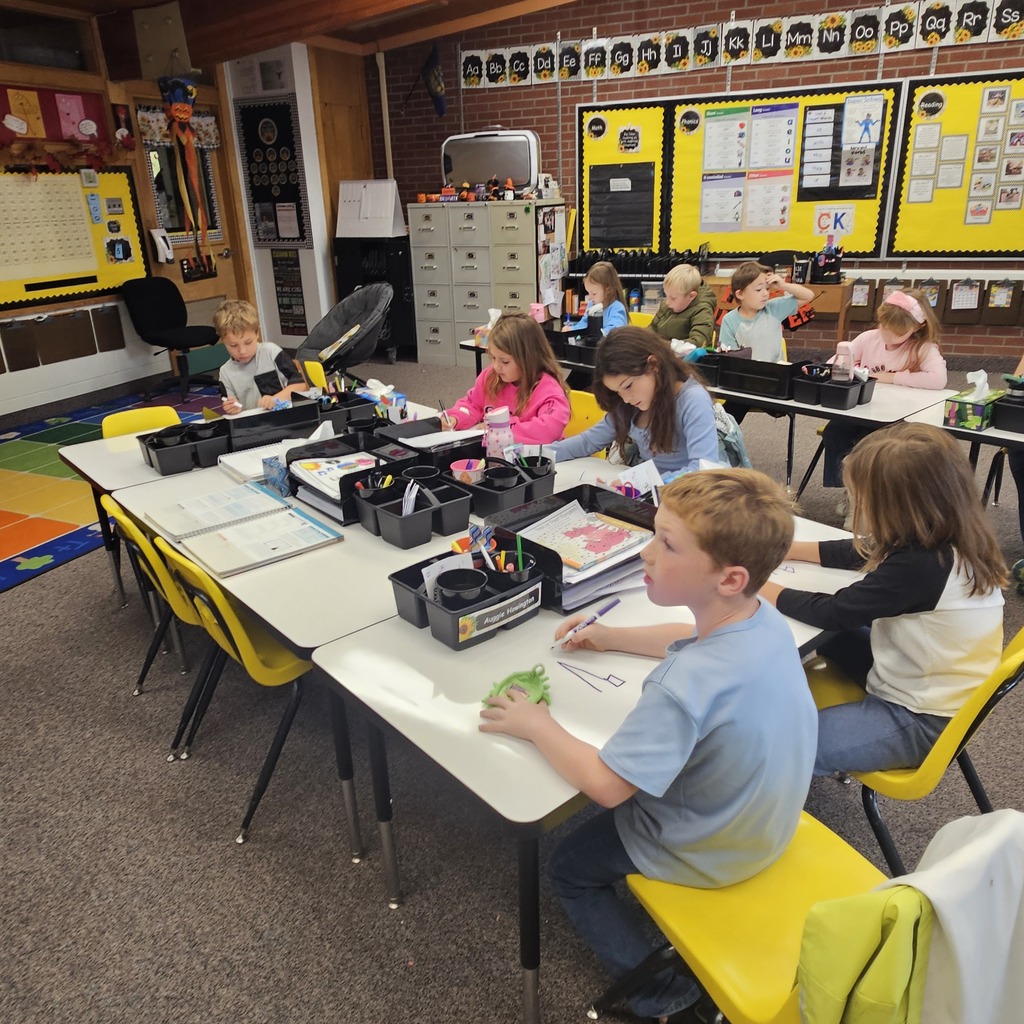 Children seated at tables in a classroom, working on a project with pens, pencils, and papers.