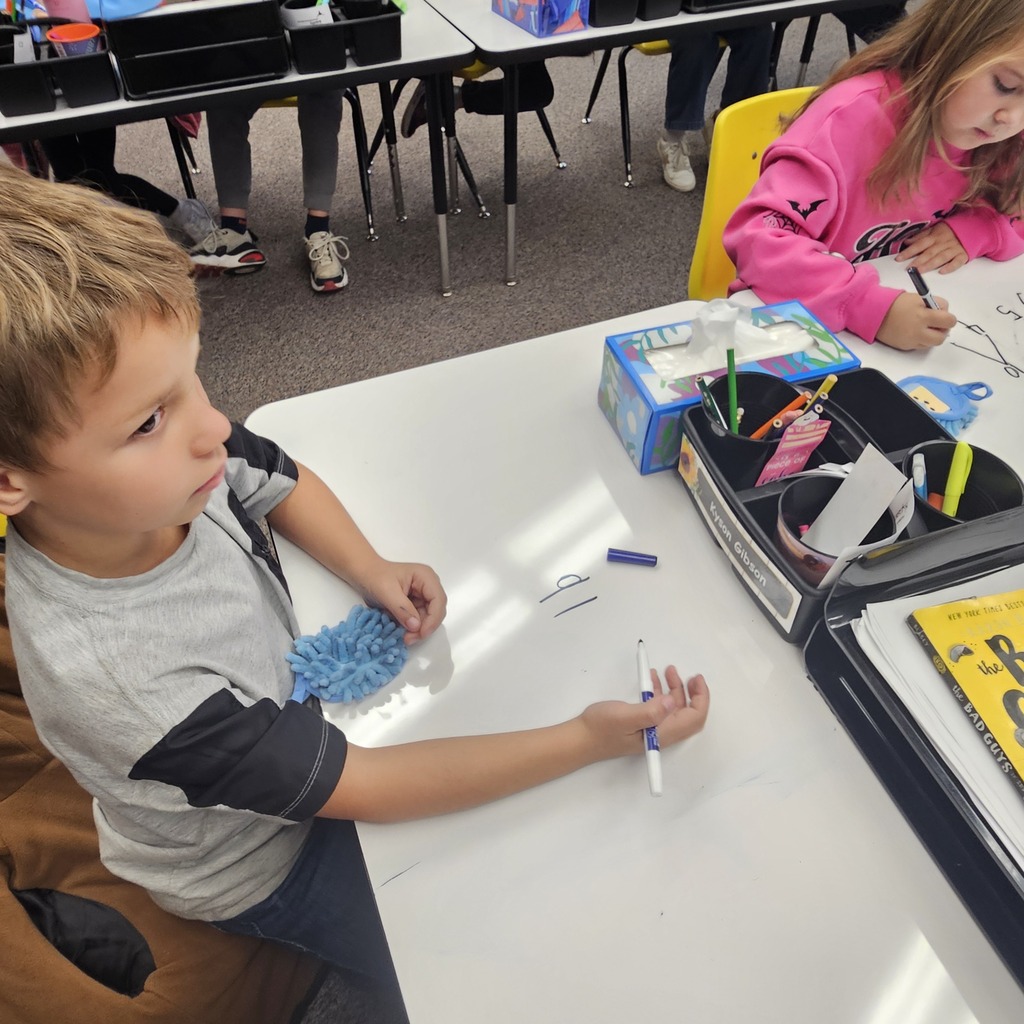 Kids seated at desks drawing with markers; one girl looking at her work and boy leaning forward.