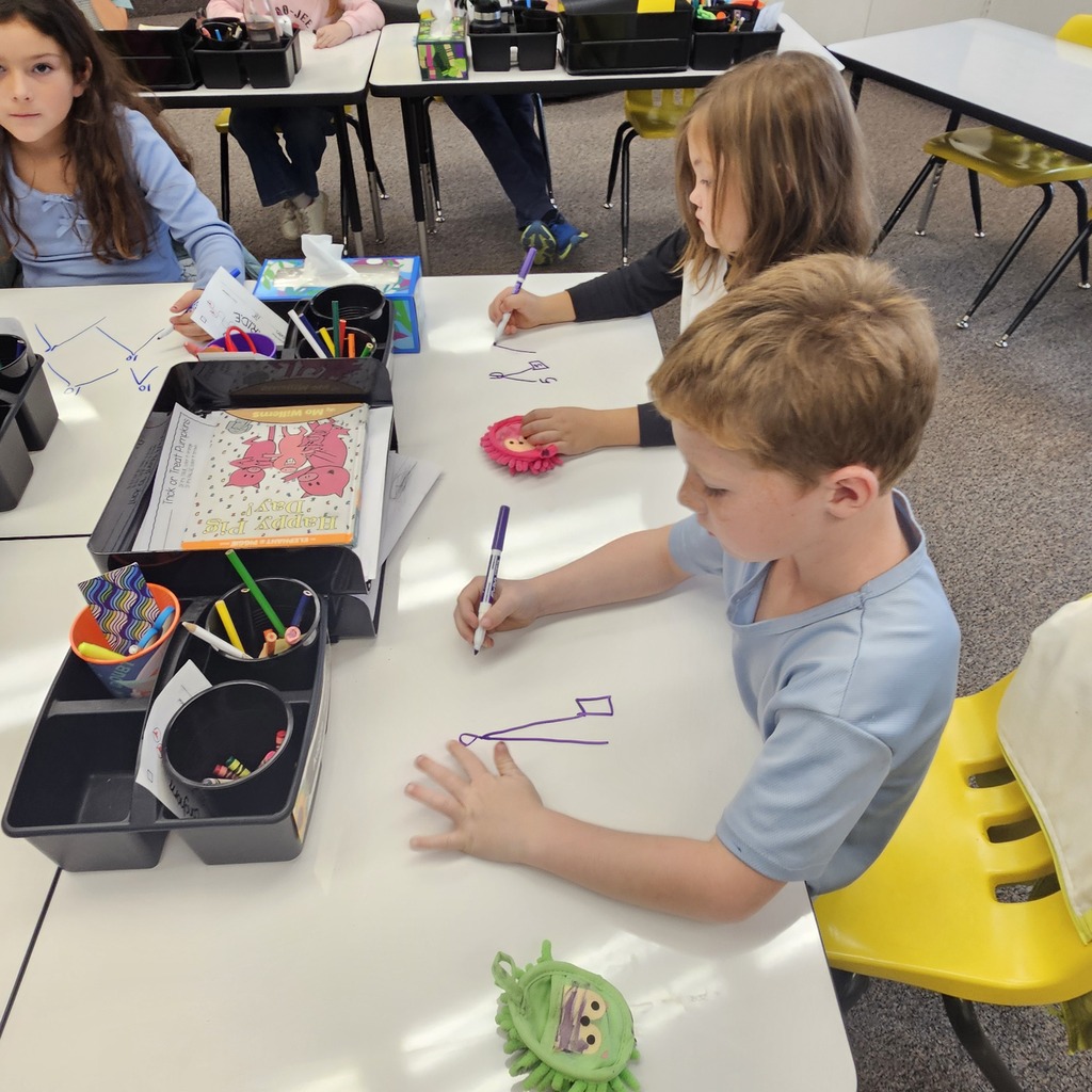 Three young children seated at a table using markers to color a large sheet of paper.