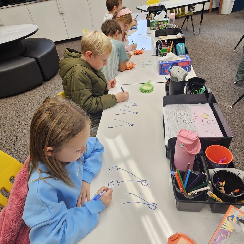 Students writing on a whiteboard with blue markers; tables with markers, cups, and paper behind them.