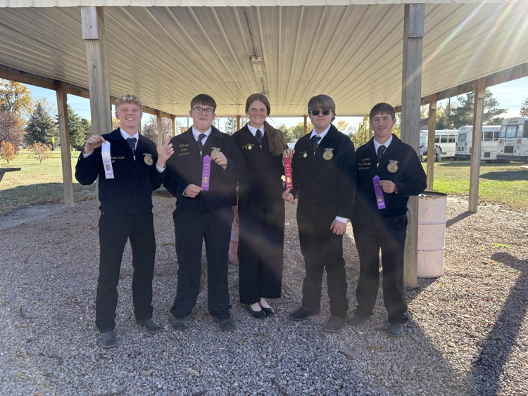 Five students in matching uniforms hold medals in front of a metal roof structure with gravel ground.