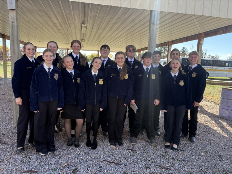 School uniform group standing on gravel under a shelter, some holding papers, with trees and a vehicle in background.