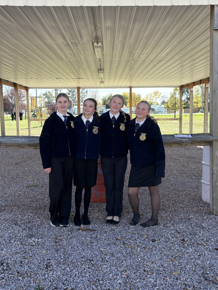 Four girls wearing school uniforms stand together in a covered outdoor area with gravel underfoot.