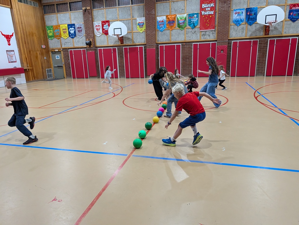 Children running and jumping over yellow and green balls on an indoor court with red walls.