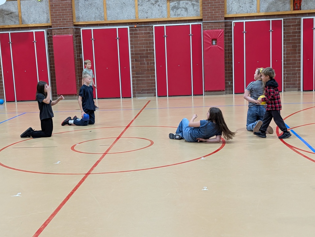 Six children in a gym with red doors and red lines on floor playing game; some kneeling, some standing