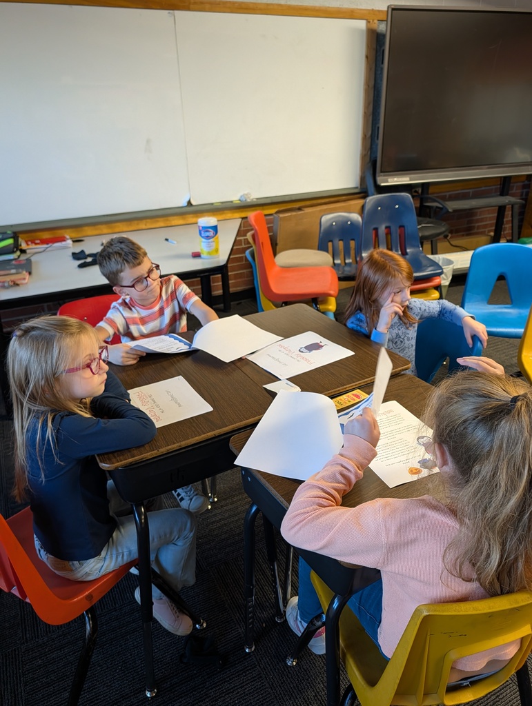 Four children seated in chairs, working at desks. One holds paper, others have books. Behind them, a whiteboard, a TV screen, and various chairs.