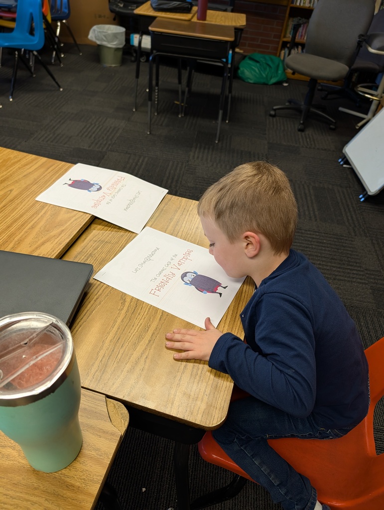 A boy is seated at a table reading a paper, with a cup nearby, in a classroom.