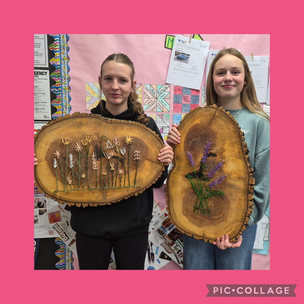 Two students stand with wood slices, one holding a slice with flowers and the other with a flower arrangement.