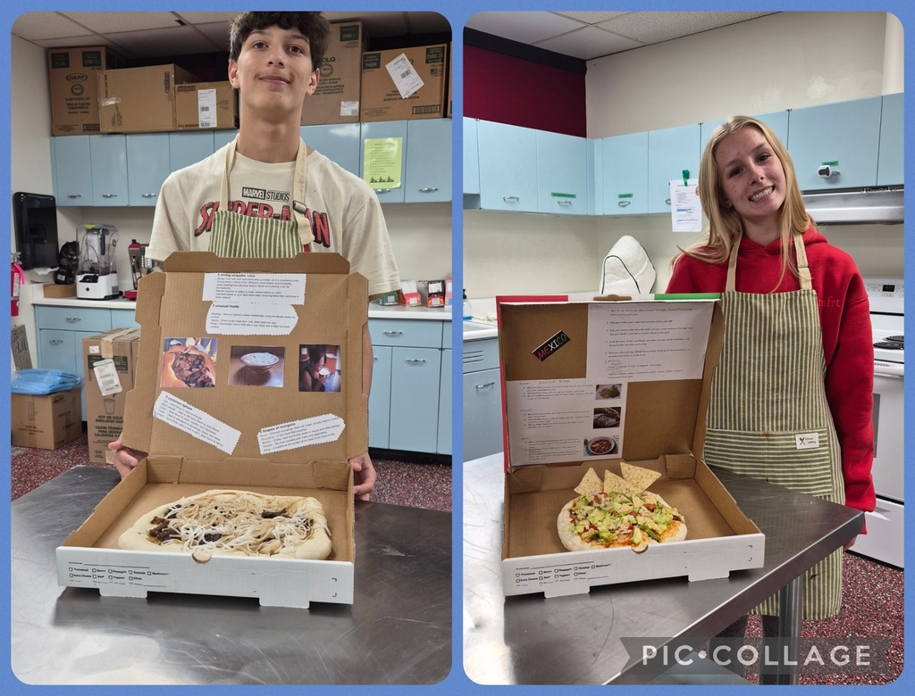 Two students, one male, one female, each hold a box displaying a pizza creation. The boy wears a beige apron, the girl a red long-sleeve shirt and green apron.