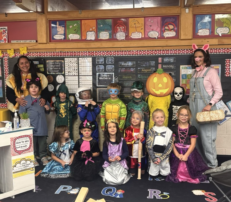 Classroom with children in costumes, one with a pumpkin, others with props, a teacher in overalls, a white counter.
