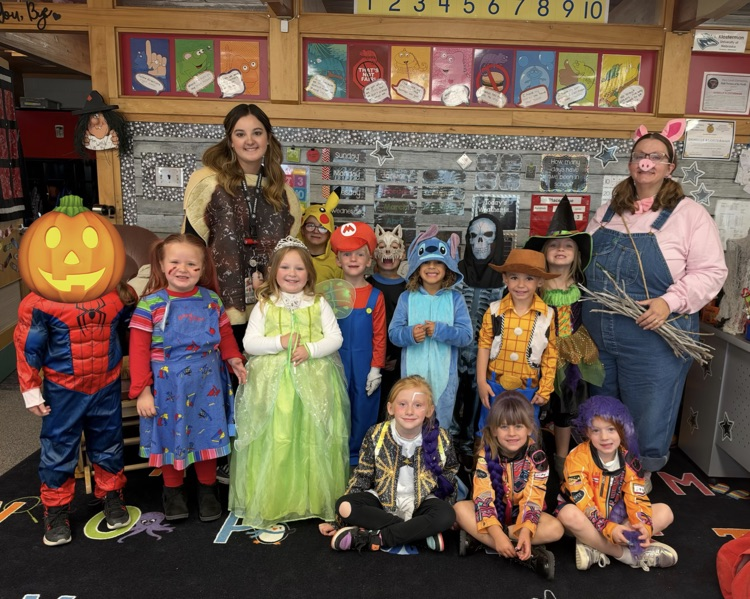 Kids dressed in costumes pose for a photo in a classroom, with letters on the floor.