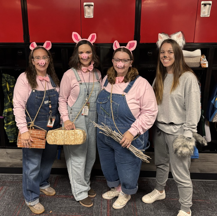 Four women in costumes stand in front of lockers, each holding an item; two have pig ears, and two wear bows.