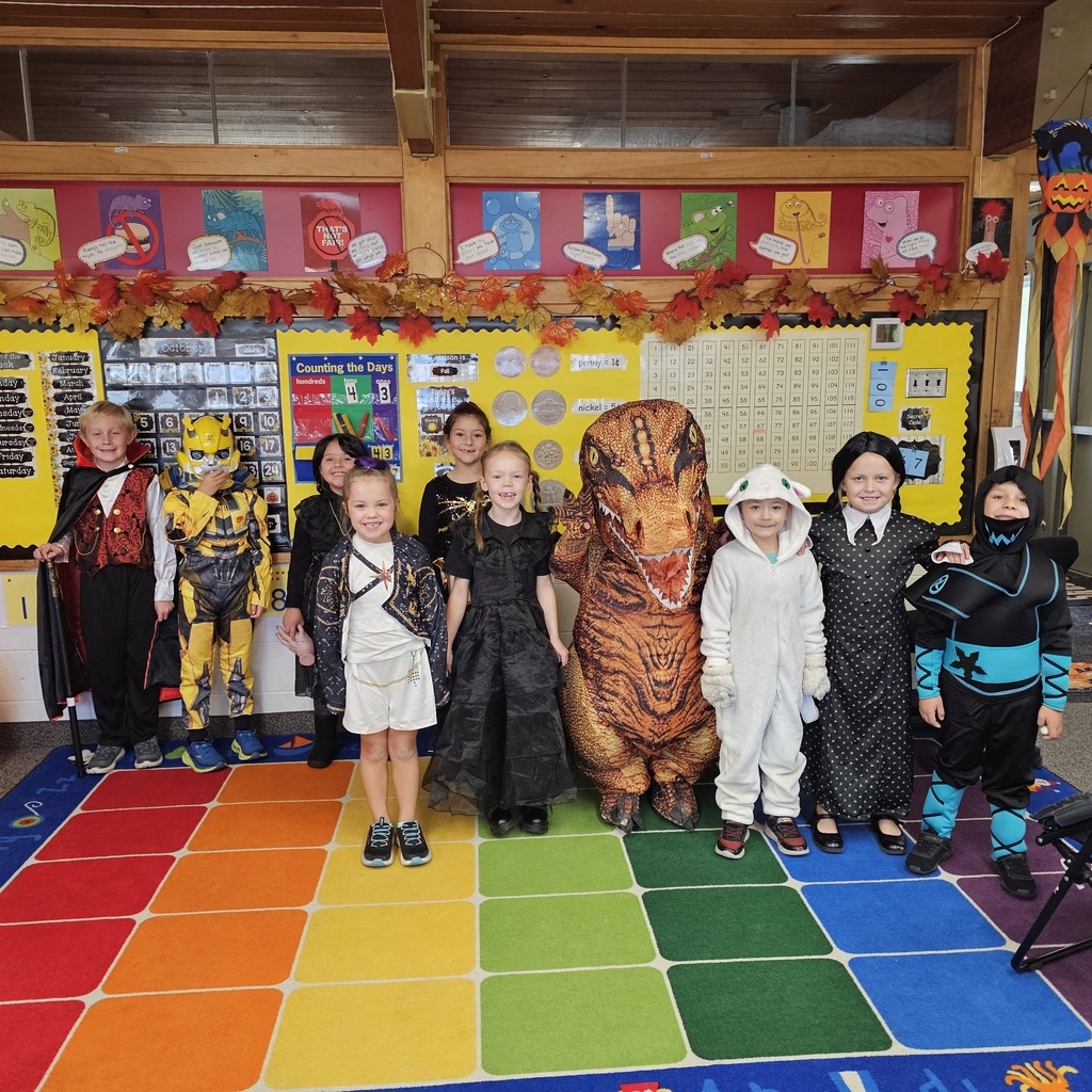 A group of children in various costumes, including a dinosaur, stand on a multicolored carpet in a classroom.