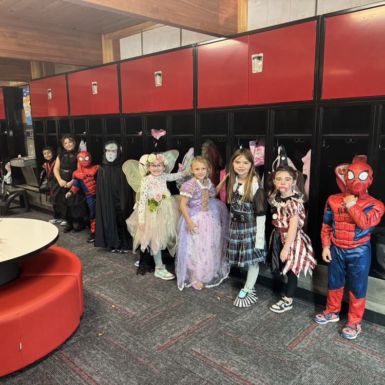 Students in costumes stand in a line, some wearing masks, in a room with lockers.