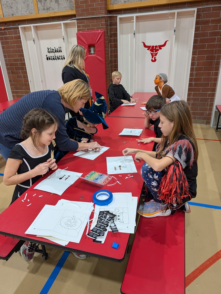 A group of children and adults are seated at a red table with papers, crafting materials, and a large red poster in the background.