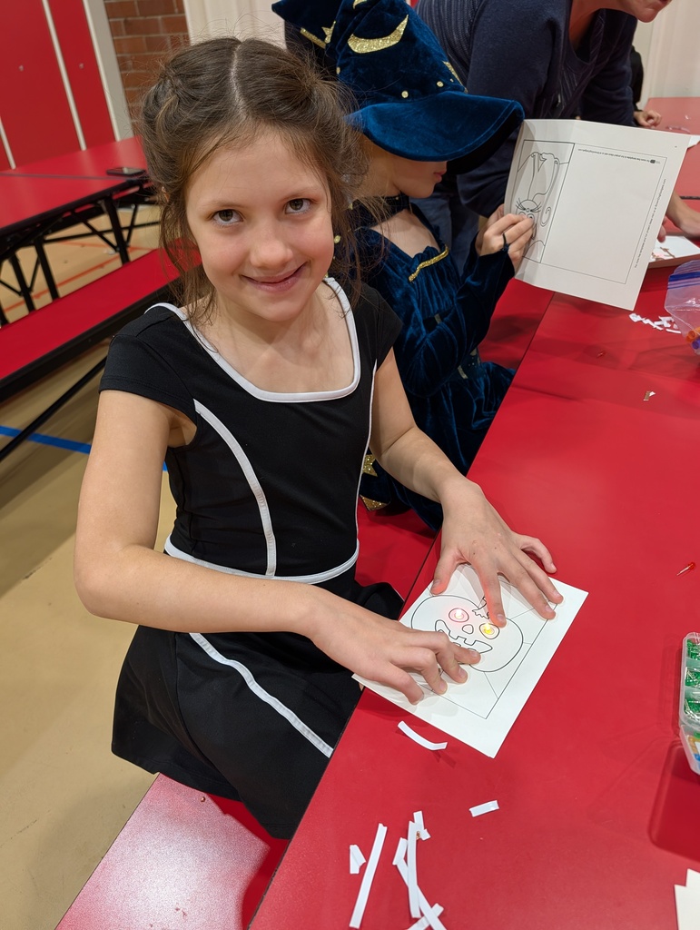 Girl sitting at a table holding a drawing with cut out shapes while a person nearby wears a hat.