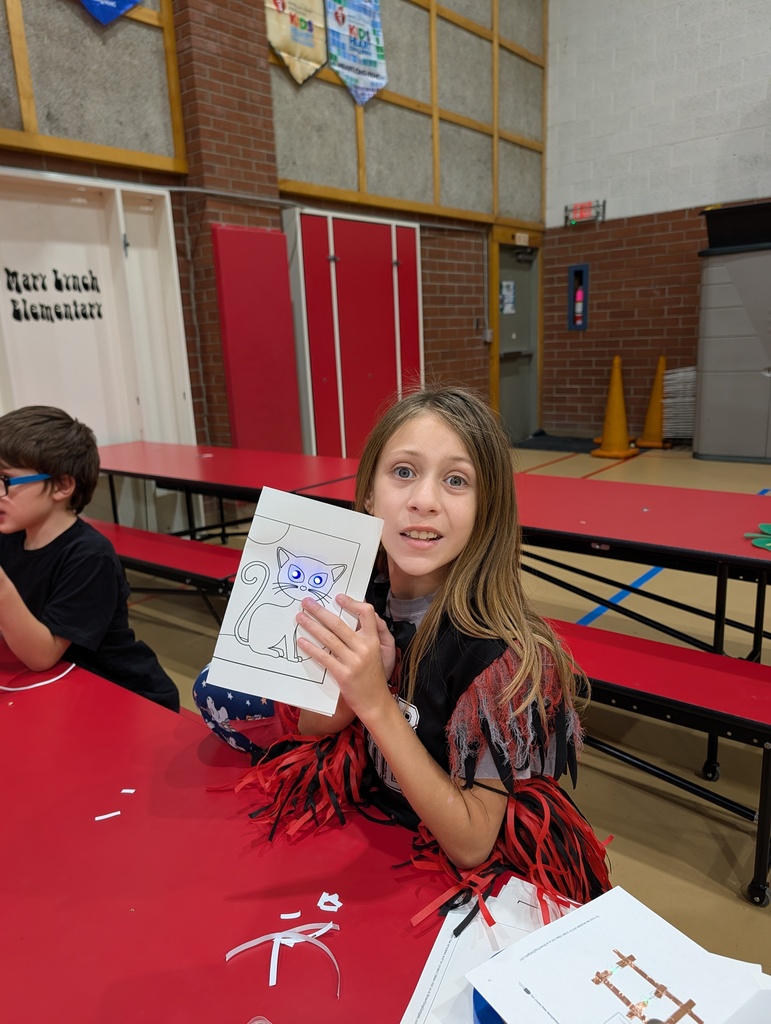 A girl smiles while holding a drawing of a cat; another child sits at the same table.