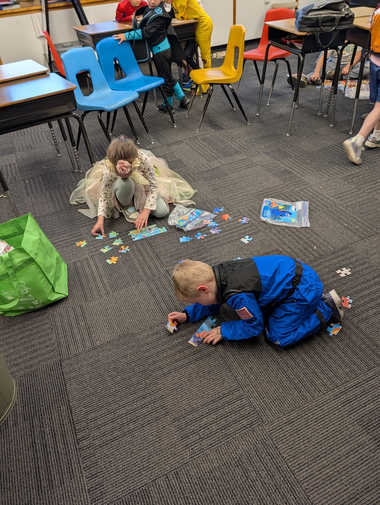 Kids sit on floor, working on puzzle pieces. Chairs, desks, and green bag in background.