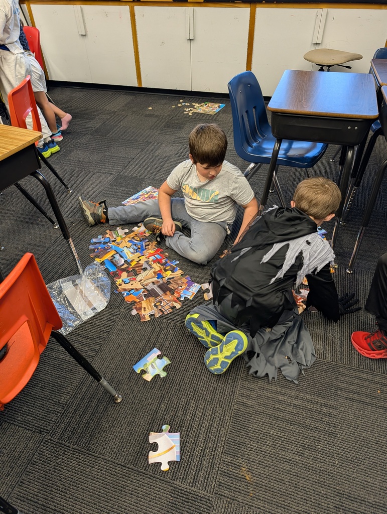 Two boys sit on the floor of a classroom, surrounded by scattered puzzle pieces. One wears sneakers; the other wears a backpack.