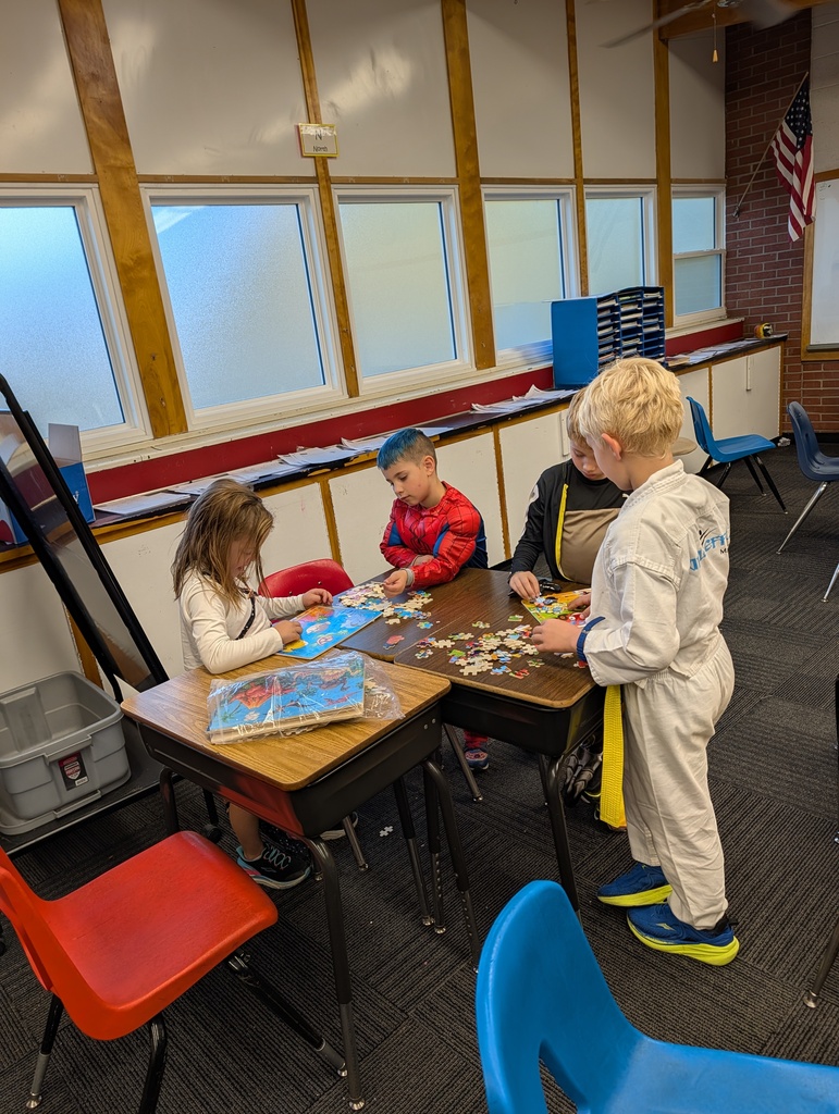 Four children work on a puzzle at desks in a classroom. A flag hangs on the wall behind them.